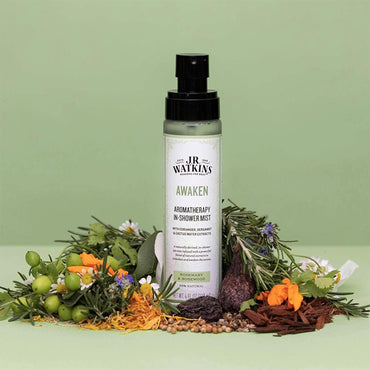 A 2 Pack of J.R. Watkins Awaken Aromatherapy in-Shower Mist Natural Rosemary & Rosewood 4 oz bottles sits amid assorted herbs, flowers, seeds, and spices on a light green background.