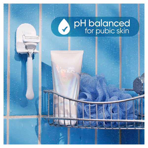 A shower scene features Gillette Venus Intimate Grooming Skin-Smoothing Exfoliant 6oz (2 Pack), a razor, and a blue bath pouf on a rack; text highlights pH-balanced formula for pubic skin and helps prevent ingrown hairs against blue tiles.