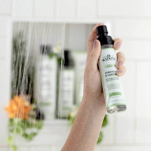 A hand holds a 2 Pack of J.R. Watkins Awaken Aromatherapy in-Shower Mist Natural Rosemary & Rosewood, with water running and blurred bottles in the background.