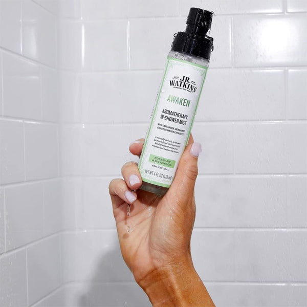 A hand holds a bottle of J.R. Watkins Awaken Aromatherapy In-Shower Mist Natural Rosemary & Rosewood (2 Pack, 4 oz) against white shower tiles, creating a spa-like atmosphere.