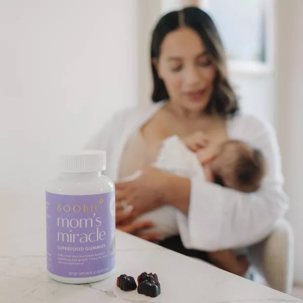 A bottle of Boobie Bears Vegan Lactation Supplement Gummies Elderberry 60ct (2 Pack) sits on a counter with two gummies in front; in the background, a woman breastfeeds, highlighting these vegan lactation support gummies.