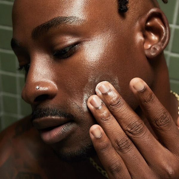 Close-up of a man applying Loved 01 Deep Moisturizing Exfoliating Cleanser (2 Pack, 6oz) to his cheek with his fingers, standing in front of a green tiled wall.