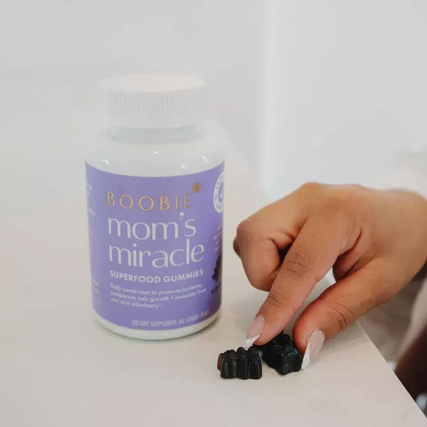 A hand picks up dark gummies next to a bottle of Boobie Bears Vegan Lactation Supplement Gummies Elderberry 60ct, a 2 pack designed to support postpartum recovery and lactation.