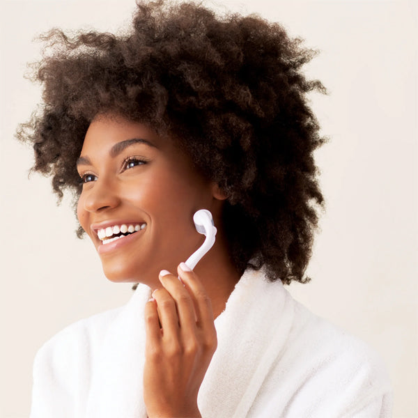 A woman in a white robe smiles while using the Plum 2 Pack - Plum Beauty Facial Microneedling Facial Roller and Massager White, shown gently massaging her cheek to promote youthful-looking skin against a neutral background.