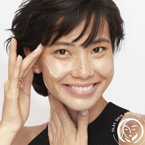 A smiling woman with short dark hair and freckles applies Olay Regenerist Collagen Peptide 24 Face Moisturizer (0.5 oz, 2 Pack) to her cheek, gently touching her neck. The Olay logo appears in the bottom right corner.