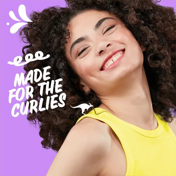 Smiling person with curly hair in a yellow top against a purple background with Made for the Curlies, celebrating Aussie Miracle Curls Shampoo and Conditioner Dual Pack 26.2 oz, enriched with coconut oil.