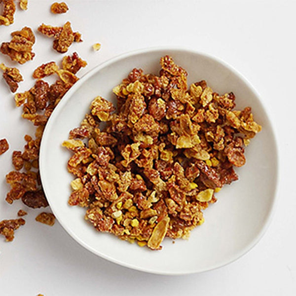 A white bowl contains granola clusters inspired by Garnier Whole Blends Repairing Honey Extracts Mask, with nourishing honey and royal jelly, scattered on a white surface next to the 2 Pack - Garnier mask.