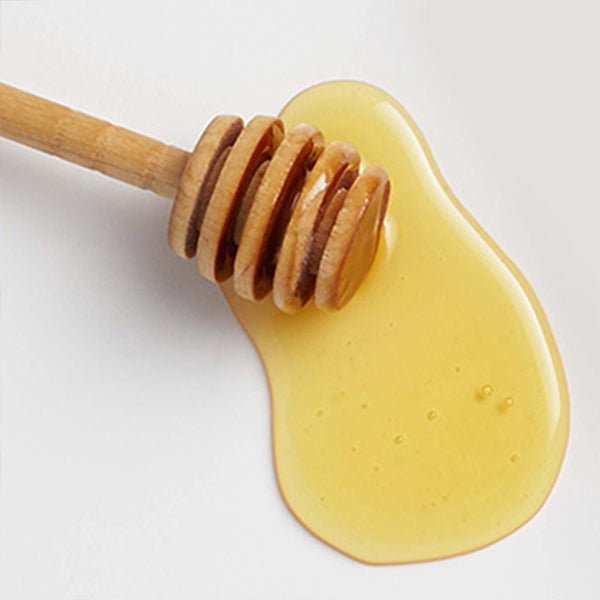 A wooden honey dipper rests in a pool of spilled golden Garnier Whole Blends Repairing Honey Extracts Mask 10.1oz, shown against a light, neutral background.