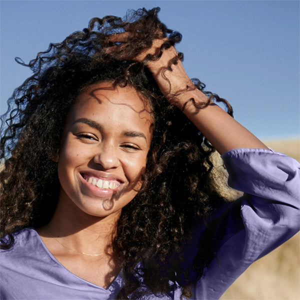 A woman with defined curls smiles at the camera in a lavender top under a clear blue sky—perhaps she used Herbal Essences Jojoba Oil & Lavender Curls Shampoo (3 Pack, 29.2oz).