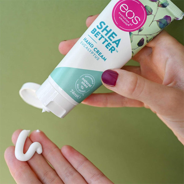 A person squeezes eos 2 Pack - EOS Eucalyptus Natural Shea Butter Hand Lotion 2.5oz onto their palm, with a promise of 24-hour hydration against a green backdrop.