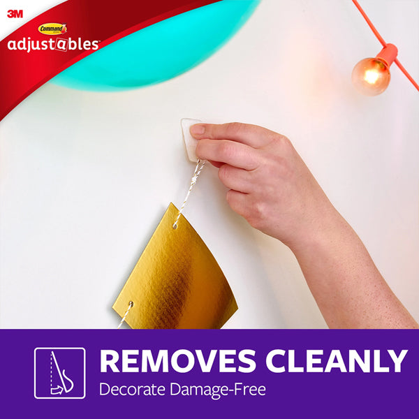 A hand removes a clear 3M 17840CLR-14ES Adjustables Clip from the wall, holding a gold decoration. Text: REMOVES CLEANLY — Decorate Damage-Free. Package shows 14 repositionable clips & 30 strips, with 3M branding visible.