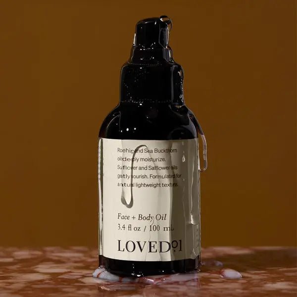 A set of three Loved 01 Face + Body Oil bottles with Rosehip & Sea Buckthorn (3.4oz each) is displayed, showcasing nourishing, plant-based hydration against a rich brown background.