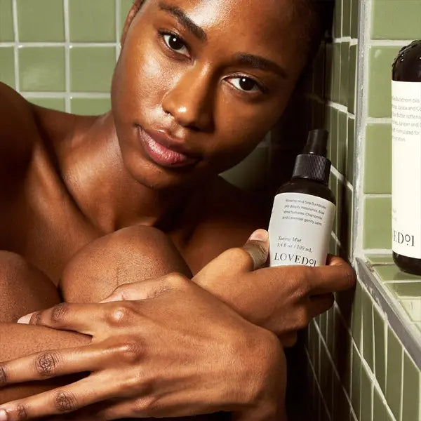 A person sits by green tile walls, holding a Loved 01 Face & Body Toning Mist Moisturizing Spray (3 Pack, 3.4oz) in one hand, with another bottle from the set placed on a nearby shelf.