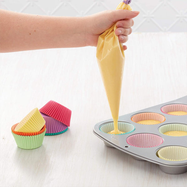 A hand uses a Wilton Disposable 16 Decorating Bag from the 4 Pack (12 Count Each) to fill colorful paper cupcake liners in a muffin tin, with extra liners stacked nearby.