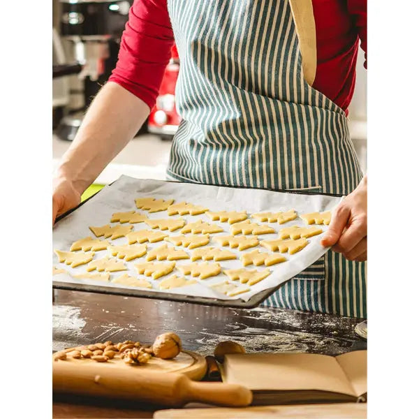 A person in a striped apron holds a baking tray lined with Kootek 12 x 16 inch parchment paper baking sheets (4 pack, 50 each), displaying rows of unbaked cookie dough in a kitchen with baking accessories and ingredients.