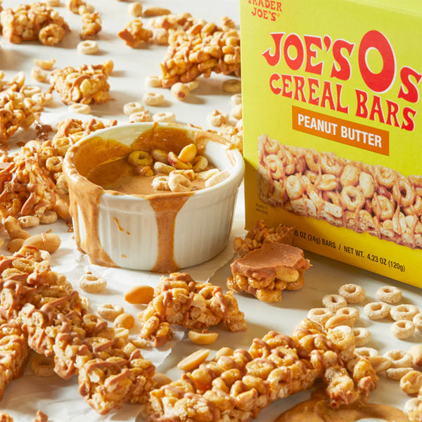 Trader Joe’s 6 Pack Joes Os Peanut Butter Cereal Bars (5 Count Each) sits by a ramekin of peanut butter, with bars, loose cereal, and peanuts—ideal breakfast snacks on a white surface.