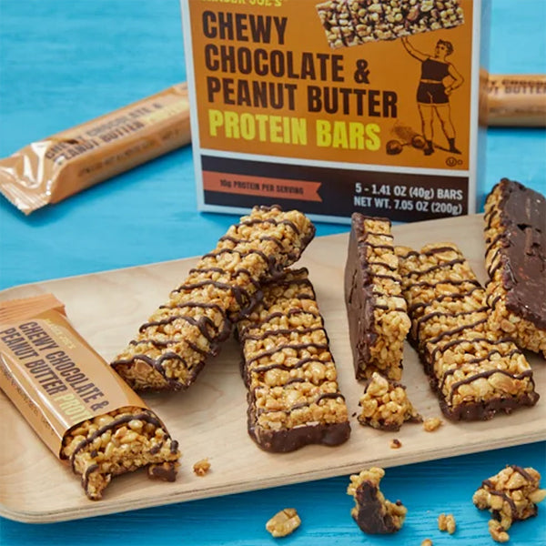 A wooden tray holds unwrapped and cut Trader Joes 6 Pack Chewy Chocolate & Peanut Butter Protein Bars (5 count each), shown with their packaging on a blue surface.