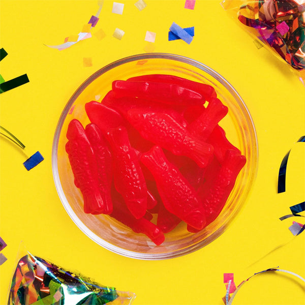 A clear bowl filled with Swedish Fish chewy candy from the 6 Pack - Swedish Fish Soft & Chewy Candy 8oz Bags sits on a yellow surface, surrounded by colorful confetti.