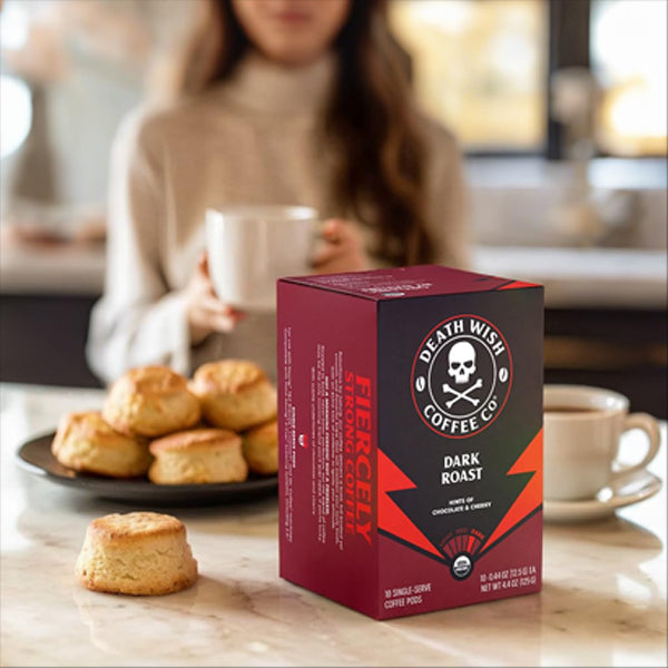 A 6 Pack of Death Wish Coffee Dark Roast Single-Serve Pods sits on a counter in front of scones, while a woman enjoys a cup of premium Death Wish coffee in the background.