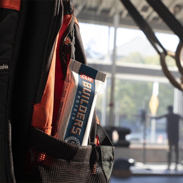 A CLIF BUILDERS Protein Bars Vanilla Almond Flavor 20g bar from Clif sits in the side pocket of a black and orange backpack, with a blurred gym background.