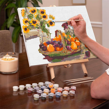 A person paints sunflowers and fruit on a Ledgebay Paint by Number for Adults Framed Canvas Country Sunshine (16x20) using paint pots and brushes, with the canvas set on an easel atop a wooden table.