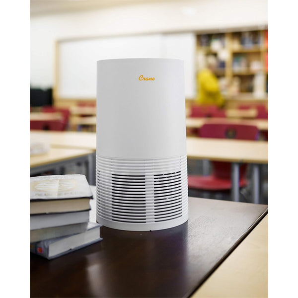 A Crane Air Purifier with True HEPA Filter and Germicidal UV Light (EE-5068) sits on a classroom table beside books, with empty desks and shelves visible in the background.