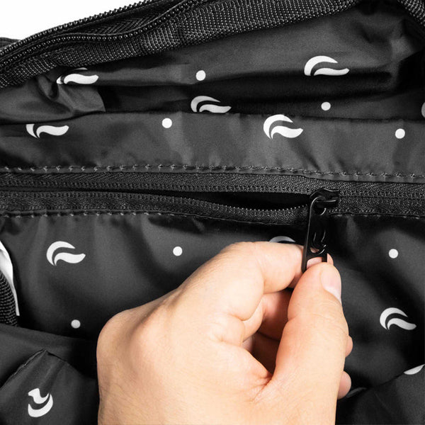 A hand unzips the Skunk Drifter Medium Crossbody Bag in Metal Gray, featuring a black zipper and a black interior with white polka dots and curved lines, plus 100% smell & weather proof carbon lining for extra freshness.