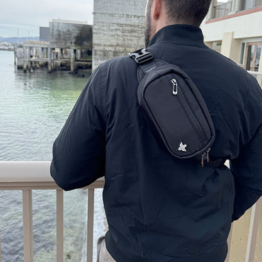 A person wearing a black jacket and the Erozul Fortuna Smell Proof Fanny Pack Crossbody Bag stands on a balcony overlooking water and distant buildings.