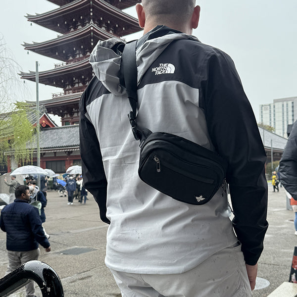 A person stands before a traditional Japanese pagoda, wearing a black and gray North Face jacket and carrying an Erozul Fortuna Smell Proof Fanny Pack Crossbody Bag, with others and umbrellas in the background.