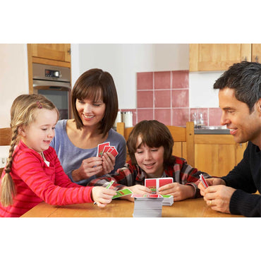 A family of four enjoys quality time at a wooden kitchen table, smiling and playing Mattel Games Apples to Apples Junior the Game of Crazy Comparisons! by Mattel—expanding their vocabulary while having fun together.
