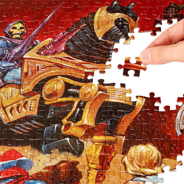 A hand completes the Mattel Games Masters of The Universe 500 Piece Jigsaw Puzzle, featuring cartoon-style armored characters and a mechanical vehicle.