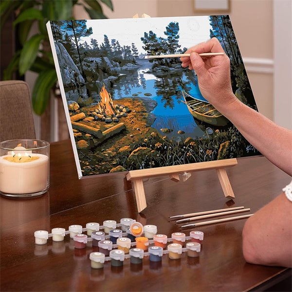 A hand paints a Ledgebay Paint by Number for Adults Framed Canvas, Moonlight Bay (16 x 20), with paint pots and brushes on a wooden table nearby.