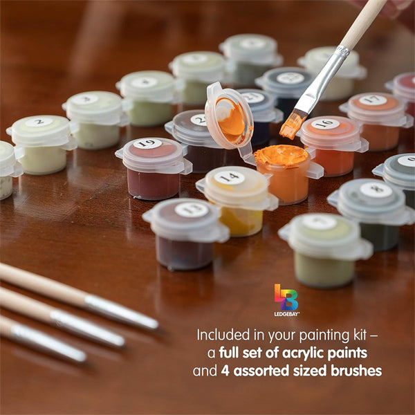 A brush dips into orange paint beside numbered containers and three brushes on wood. Text highlights that the Ledgebay Paint by Number for Adults Framed Canvas Moonlight Bay, 16 x 20, includes all essentials for a DIY project.