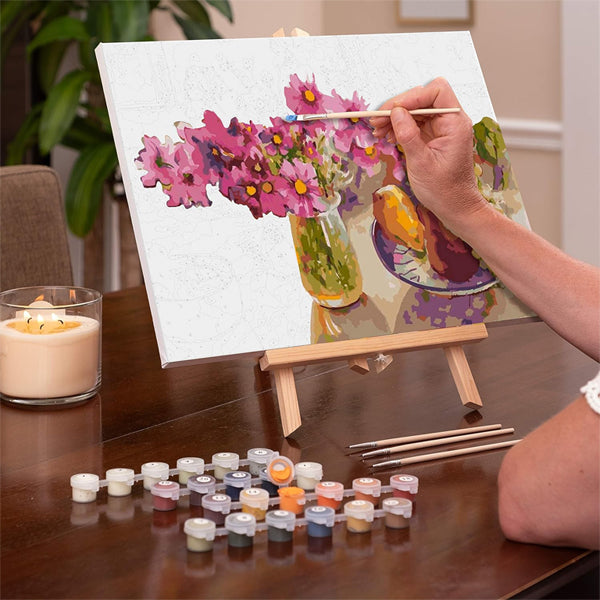 An adult enjoys painting Cosmos from The Garden on a 12x16 framed canvas using the Ledgebay Paint by Number kit, with paint pots and brushes arranged nearby on a wooden table.