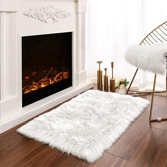 The Latepis White Luxury Faux Fur Sheepskin Rug 2x3 is placed on a wooden floor near a lit fireplace, accompanied by a gold chair and elegant decor items.