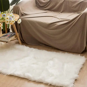 A beige couch with a checkered cover sits behind a small wooden side table topped with flowers and books; the Latepis White Luxury Faux Fur Sheepskin Rug 2x3 adds plush comfort to the wooden floor in front.