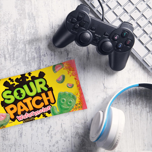 A 24-count box of Sour Patch Kids Watermelon Soft & Chewy Candy 2oz bags sits beside a game controller, white keyboard, and white headphones with blue accents—ideal for snacking on during gaming sessions.