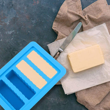 A block of butter on wax paper sits beside the Deal Society Gourmet Silicone Butter Mold with Lid, a rectangle tray with 4 cavities, and a knife on a brown cloth, all arranged on a dark surface.