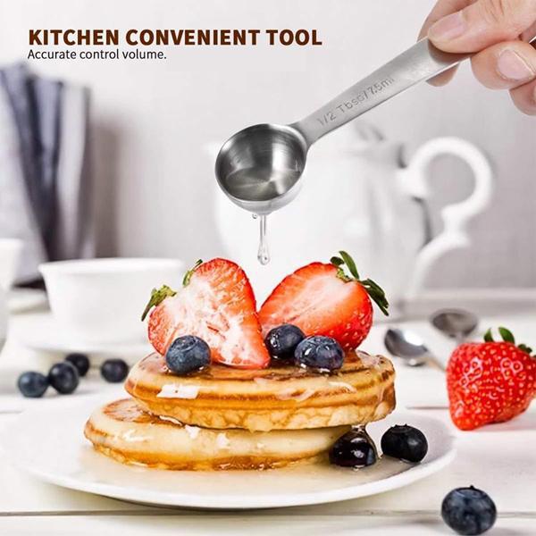 A hand uses Deal Societys Stainless Steel Measuring Spoons with Ring Holder (Set of 9) to drizzle liquid over pancakes topped with strawberries and blueberries on a white table setting.
