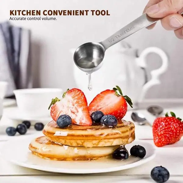 A hand uses Deal Societys Stainless Steel Measuring Spoons with Ring Holder (Set of 9) to drizzle liquid over pancakes topped with strawberries and blueberries on a white table setting.