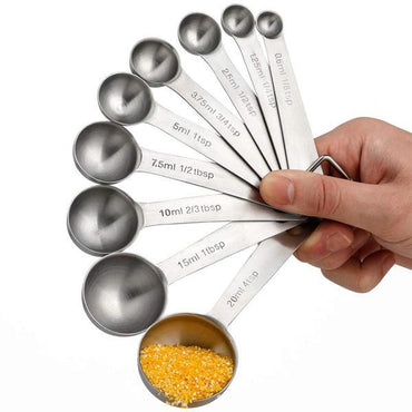 A hand holds Deal Society’s Stainless Steel Measuring Spoons with Ring Holder, set of 9, each with engraved markings; the largest spoon contains yellow cornmeal.