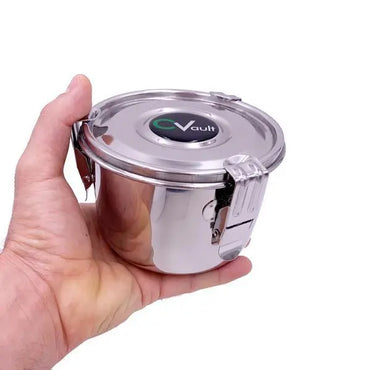 A hand holds a CVault Large Humidity Control Airtight Metal Smell Proof Container by CVault, featuring a round stainless steel body, secure metal latch, and a lid labeled “CVault” against a plain white background.