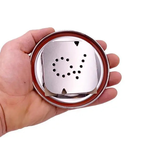 A hand holds a CVault Medium Humidity Control Airtight Metal Stash Smell Proof Container by CVault, featuring a round metal body and a square perforated plate with a spiral hole pattern.