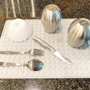 Stainless steel cups, a white bowl, and cutlery are placed on the Deal Society Self Draining Silicone Dish Drying Mat 16 x 12 Twin Pack for fast drying on the kitchen countertop.