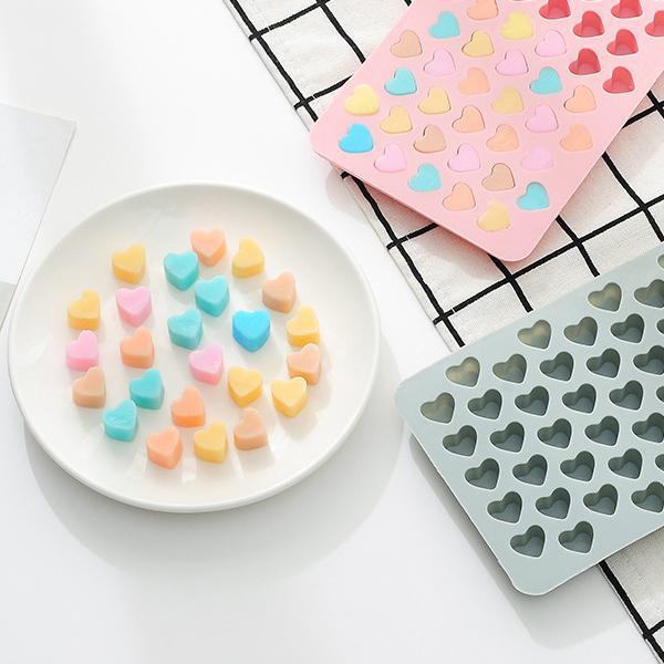 A white plate with colorful heart-shaped candies sits beside two Deal Society Silicone Gummy Heart Molds, included in the 3 pack, one pink and one gray, displayed on a white surface with a checkered cloth.