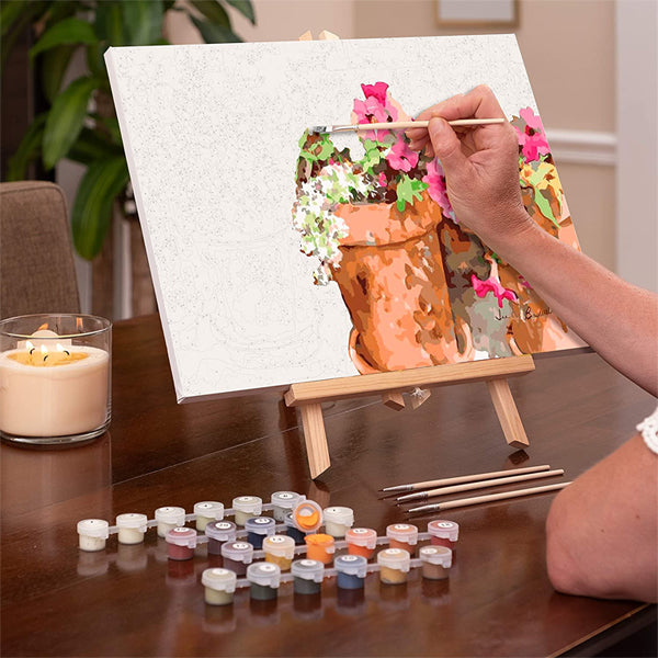 Using the Ledgebay Paint by Numbers Kit for Adults – Sweet Petunias (16 x 20) framed painting, someone paints floral terracotta pots at an easel with paint pots, brushes, and a candle arranged on a wooden table.