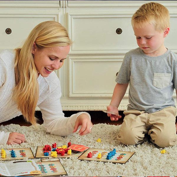 A woman and a young boy sit on a carpeted floor playing Inspiration Play Rail Wreck, a colorful train-themed educational board game for toddlers and preschoolers, in a bright room.