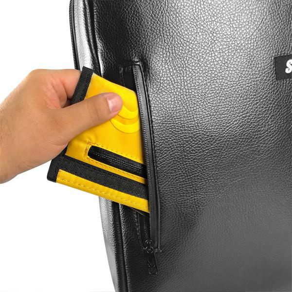 A hand places a yellow and black Skunk Elite Backpack Smell Proof Weather Proof Back Pack with combo lock—100% odor proof—into the zippered pocket of a black leather bag.