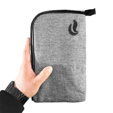 A hand holds the Skunk Travel Pack 10 Smell Proof Bag—a gray fabric pouch with a black logo, wrist strap, and lockable zipper for added security by Skunk.