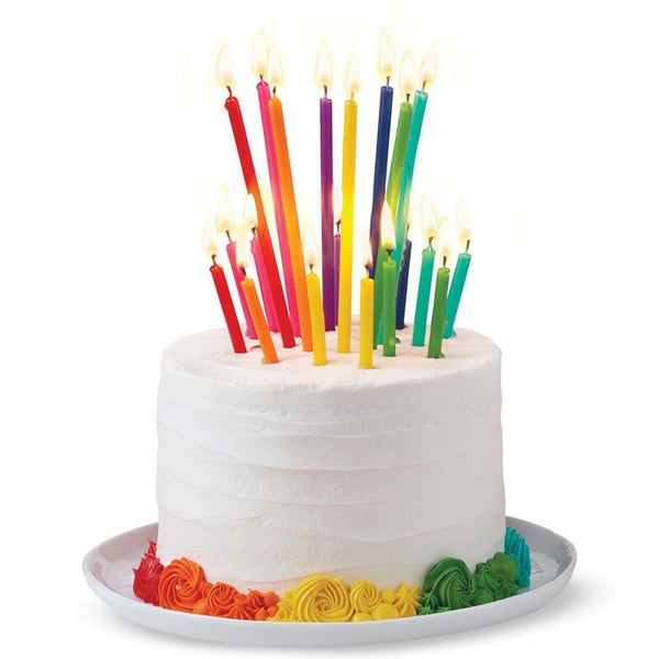 A white frosted cake sits on a white plate, decorated with rainbow icing and topped with Wilton Birthday Candles from the 5 Pack—Tall & Short set (20 count each)—all lit and colorful in various heights.